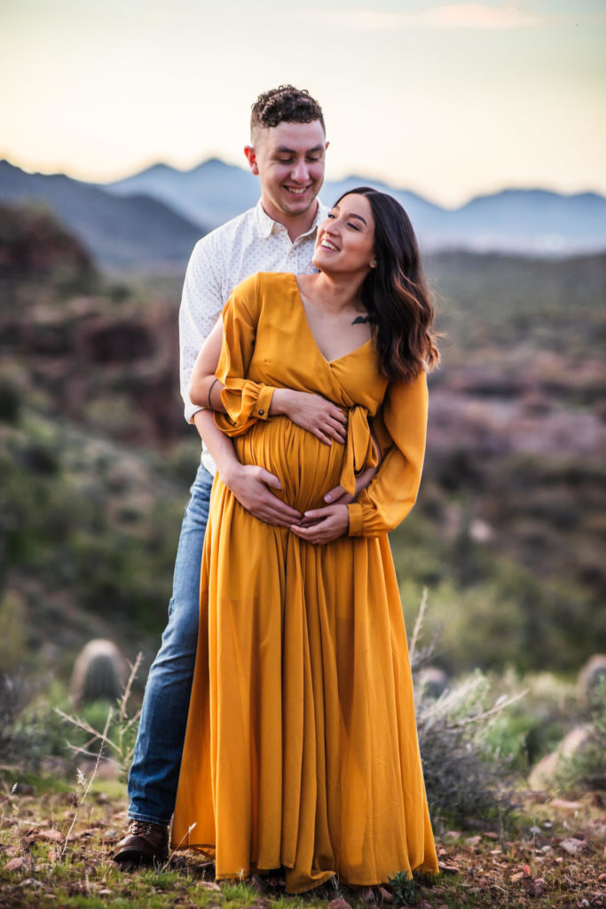 Maternity Photography in Mesa AZ - A pregnant mother stands close in front of her husband as they hug her belly during a maternity photo session.